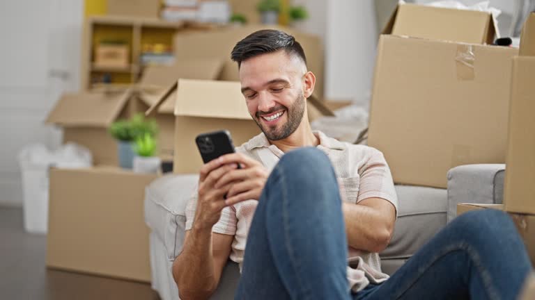 Young hispanic man using smartphone sitting on floor at new home