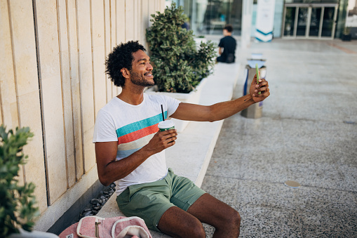 Black man enjoying a day spent alone sitting on a park bench, drinking coffee from a disposable cup and taking a selfie photo