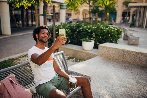 Multiracial man enjoys spending time alone outdoors. He is sitting on a park bench and taking a selfie
