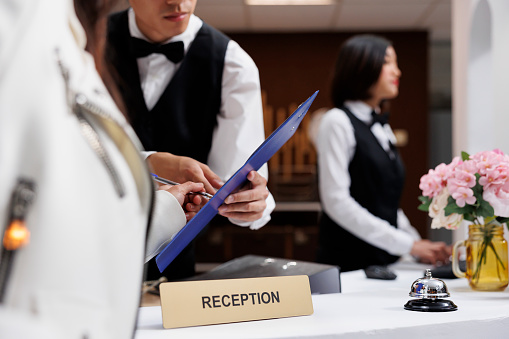 Male Asian receptionist at front desk of contemporary hotel assists customer in signing the registration form. Woman goes on vacation and checks into welcoming resort in foyer