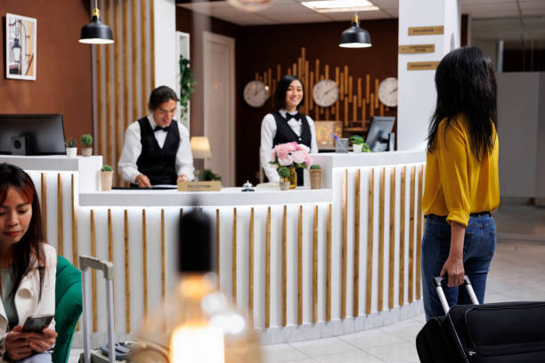 Vacationers arrive at inviting hotel entrance with baggages and are waiting to make room reservations. Stylish hotel lobby with fresh guests and two smiling Asian receptionists