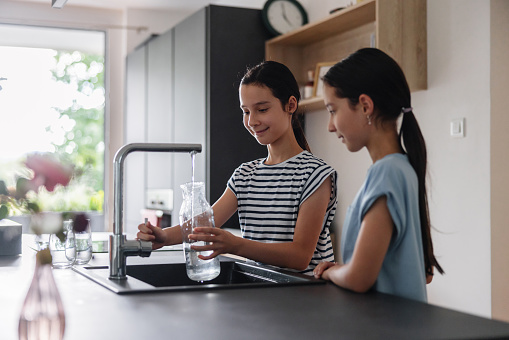 Two young girls wearing casual clothes and ponytails are standing in front of the sink while one of them is filling a glass jar with some tap water. They are at a modern kitchen with daylight.