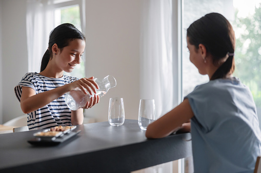 Two multiracial young sisters are together at a table, while one of them is holding a glass jar and preparing to serve some water in one glass. The sister is sitting in front of her and observing how she does it.