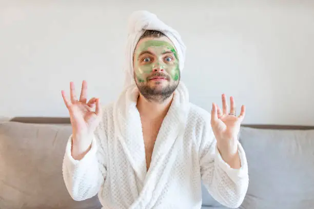 Photo of Funny man enjoys beauty treatments and luxury relaxation at home spa. A man with a towel on his head sits on a sofa in a white bathrobe and with a cosmetic mask on his face.