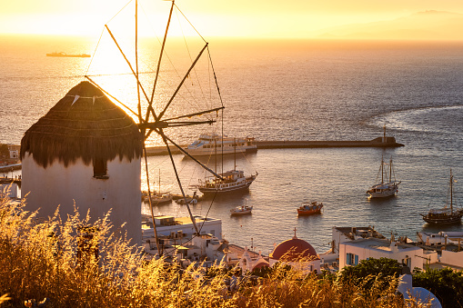 Golden sunset over sea horizon in Mykonos, Cyclades, Greece. Famous traditional white windmill overlook civil port and harbor on hot summer evening in bright sunlight. Romantic image, iconic travel destinations