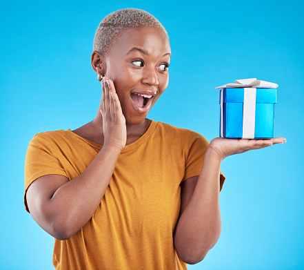 Wow, gift and excited black woman with box in studio isolated on a blue background. Surprise, happy and African person with present for party, celebration of holiday or birthday package for giveaway