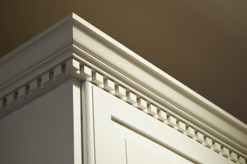 The top crown moulding dentil detail on some creme-color painted solid wood kitchen cabinets. These wall mounted cabinets do not go all the way to the ceiling above them. The lighting is coming from the top right from a ceiling light.