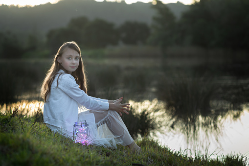 Back lit portrait of a young girl dressed in white sitting at the water's edge looking at the camera