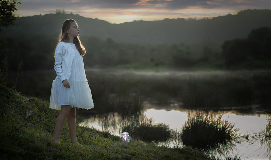 Back lit portrait of a young girl dressed in white standing at the water's edge at sunset