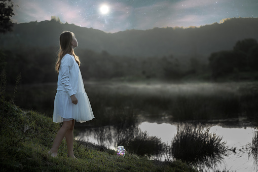 Back lit portrait of a young girl dressed in white standing at the water's edge at dusk looking at the moon rising over the mountain