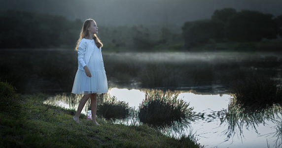 Back lit portrait of a young girl dressed in white standing at the water's edge at dusk