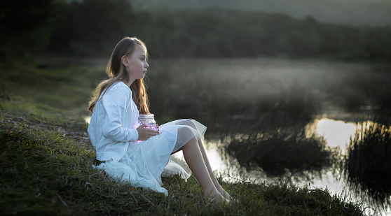 Portrait of a young girl holding a lantern sitting at the water's edge covered in a soft mist at dusk