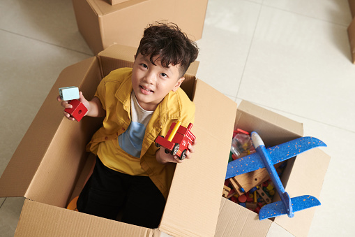 Preteen boy sitting in cardboard box and playing with toys, view from above