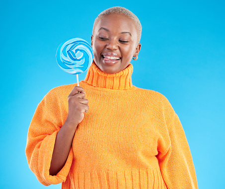 Smile, lollipop and black woman with candy in studio isolated on a blue background. African person, happy and tongue out for sweets, dessert and treats, sugar food and confectionary for eating