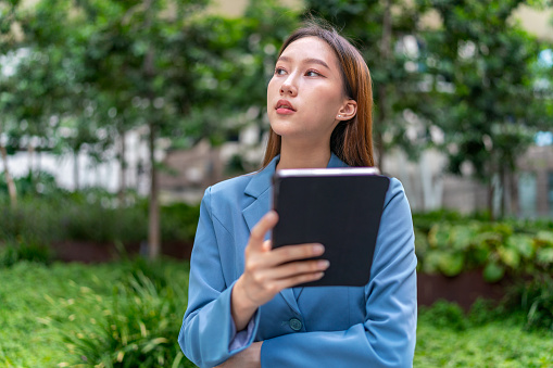 Amid the corporate cityscape, the young Asian businesswoman harnesses efficiency, working on her digital tablet in front of contemporary buildings, embodying the spirit of modern business on the move.