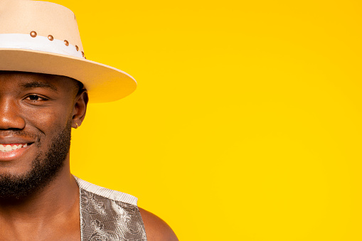 Half face of a young african happy smiling bearded handsome man wears hat and vest at studio isolated over yellow background. Fashion portrait.