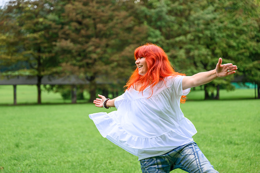 Cheerful senior woman with long dyed red or orange hair, wearing jeans and white blouse, walking, running or dancing on a green meadow in a public park.