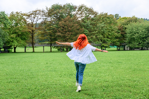 Cheerful senior woman with long dyed red or orange hair, wearing jeans and white blouse, walking, running or dancing on a green meadow in a public park.