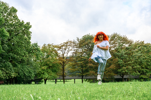 Cheerful senior woman with long dyed red or orange hair, wearing jeans and white blouse, walking, running or dancing on a green meadow in a public park.