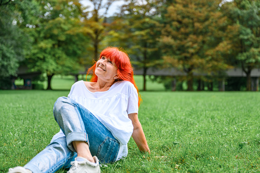 Senior woman with long dyed orange hair, wearing jeans and white blouse sitting on a green grass in a public park.