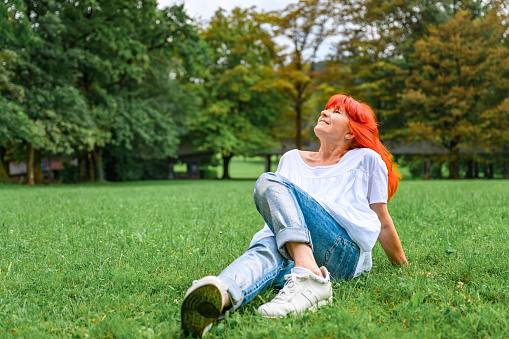 Senior woman with long dyed orange hair, wearing jeans and white blouse sitting on a green grass in a public park.
