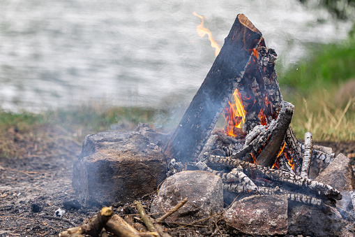 Close up of burning brushwood campfire on forest ground on blurred background of river, camp fire in cloudy day outdoors.