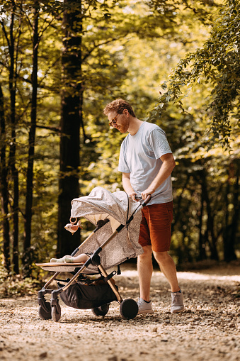 A young white man walking in the park with his newborn baby, pushing the baby pram along an unpaved pathway.
