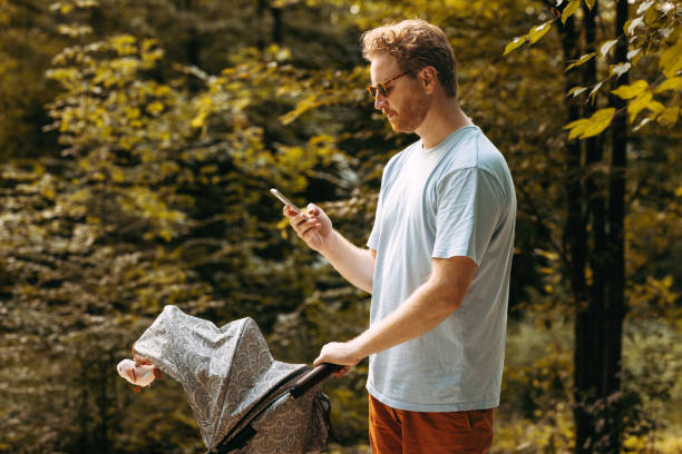 A young white father using his mobile phone while walking in the park with his baby in a pram.