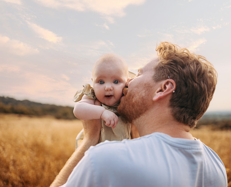 A portrait of a happy white man giving his baby a kiss on the cheek as they walk in the fields.