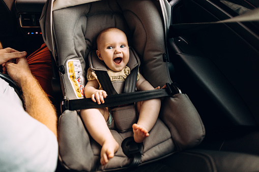 A happy Caucasian newborn baby sitting in an infant carrier car seat, next to her unrecognizable father.