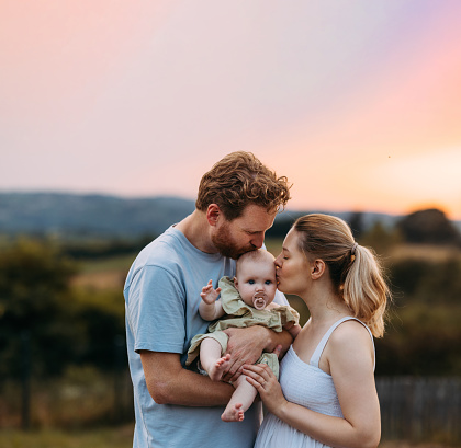 A portrait of happy Caucasian parents holding their infant and giving her a kiss in the countryside.