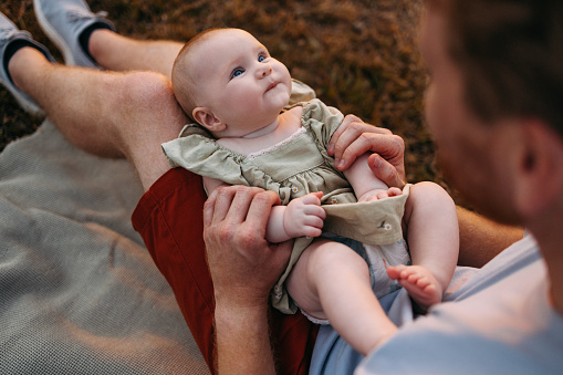 An anonymous white man sitting on a blanket and holding his smiling newborn child on his lap, playing with her.