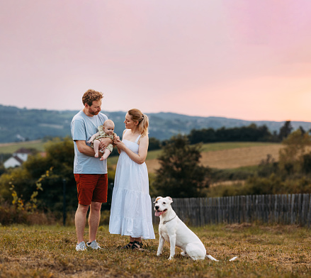 A portrait of a happy mother and father standing in their backyard, holding their newborn baby, with a  white dog sitting by their side.