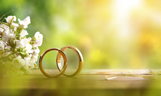 Detail of two gold rings on a rustic wooden table with flower bouquet outside with nature background. Front view.