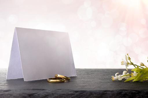 Two engagement rings on slate table with blank paper sign behind and flower bouquet with light pink background and bokeh effect. Front view.