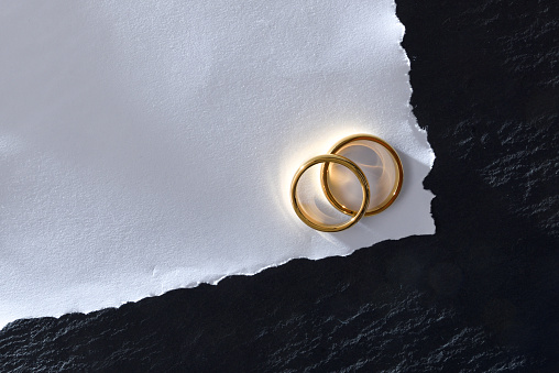Macro of white textured paper card with two engagement rings on slate base table. Top view.