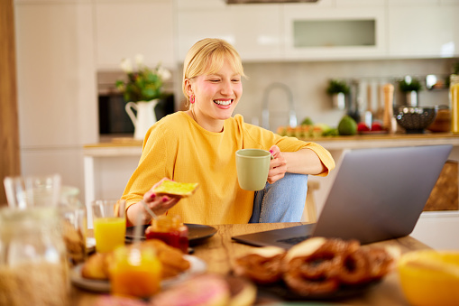 Young woman drinking coffee, eating and working on her laptop over breakfast at home