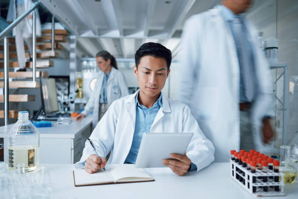 homem asiático, comprimido e escrita em laboratório de ciência para pesquisa de vírus médicos, medicina e desenvolvimento de vacinas. cientista ocupado, tecnologia e papel de caderno para amostra de sangue saúde ou bem-estar - medical research healthy lifestyle paper healthcare and medicine - fotografias e filmes do acervo