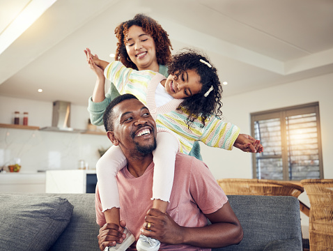 Black family, father and daughter playing with mother on living room sofa together for fun bonding at home. Happy dad carrying child for piggyback ride with mom on lounge couch enjoying holiday break