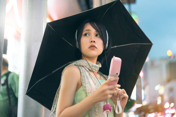 Asian woman wear nostalgia stylish using headphone and flip phone with umbrella on rainy season at night city Urban real people lifestyles in downtown concept. Young asian woman wear y2k nostalgia trend outfit stylish using headphone and flip phone. Rainy season with umbrella. Stand in Bangkok city Thailand person on flip phone stock pictures, royalty-free photos & images