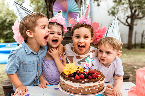 Cute little girl celebrating 3rd birthday with friends in backyard, blowing birthday cake candles.