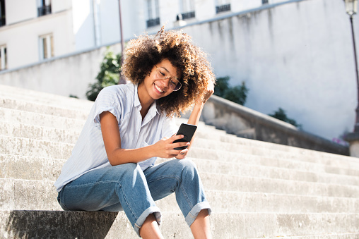 Portrait smiling young woman with eyeglasses sitting outside looking at phone