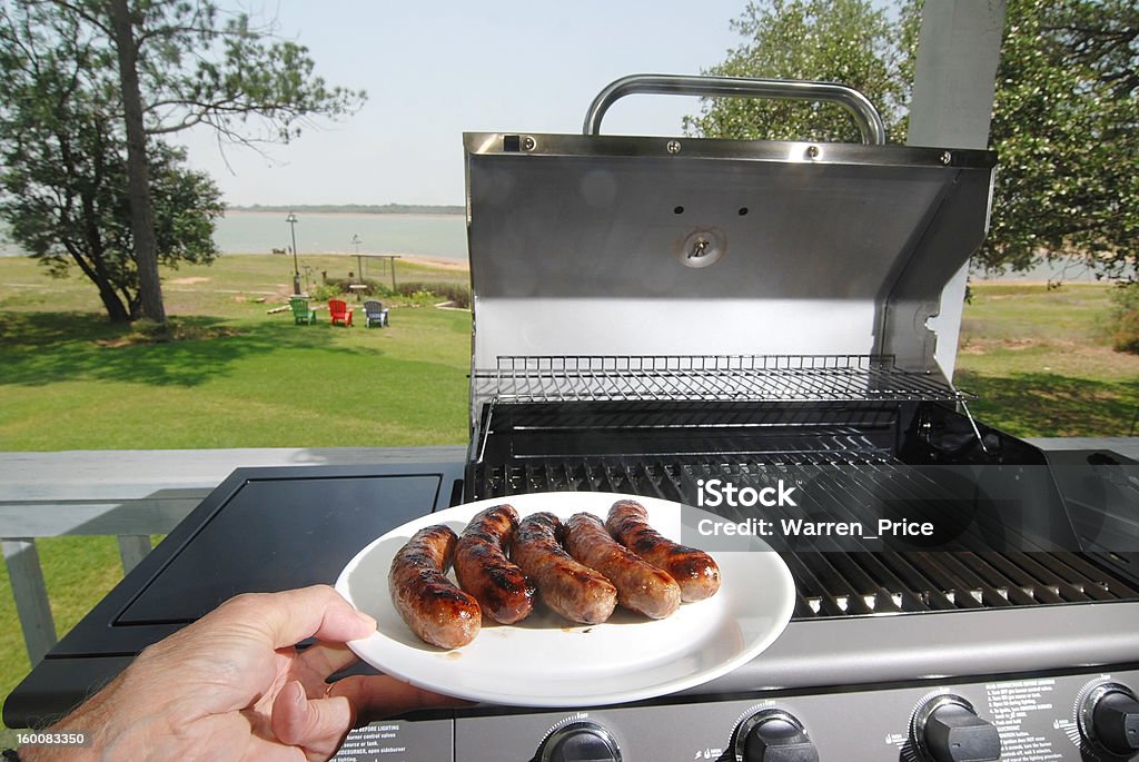 Summer Holidays Cookout Hand holding plate of sausages just taken from the grill with lake and green lawn as background. Barbecue Grill Stock Photo