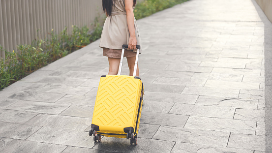 Freelance young adult woman walking with travel luggage for business trip. Rear view of female long hair casual beige suit. Urban lifestyles people on day.