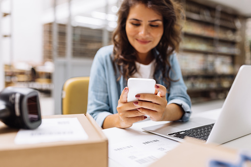 Online store manager sending a text message on her cellphone. Creative businesswoman making plans for product shipping in her warehouse. Young female entrepreneur running an e-commerce small business.