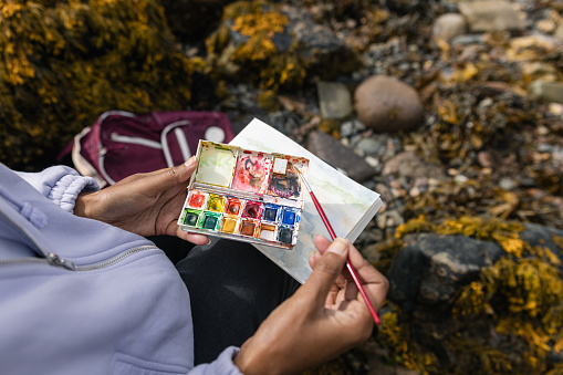 An over the shoulder view of an unrecognisable mature woman sitting on the rocks at the waters edge at a beach in Torridon, Scotland. She is painting in a book with a paintbrush while enjoying her surroundings.