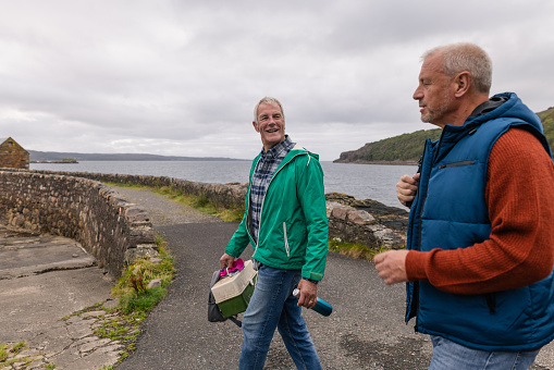 A side view of a mature and senior man walking together near the coastline, they are they are carrying a lunch box and before they take the boat out on the Scottish waters in Torridon, Scotland.