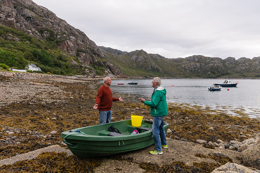 A wide side view of a mature and senior man standing together talking at the waters edge while preparing their rowing boat. They are nearly ready to take the boat out on the Scottish waters in Torridon, Scotland.