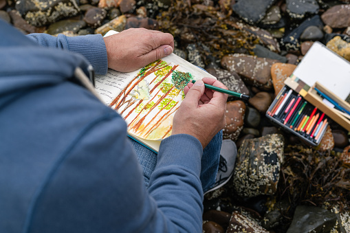 An over the shoulder view of an unrecognisable mature man sitting on the rocks at the waters edge at a beach in Torridon, Scotland. He is drawing in a book with colourful pencils while enjoying his surroundings.