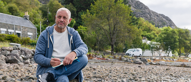 A wide view of a mature man sitting on the rocks at the waters edge at a beach in Torridon, Scotland. He is painting in a book with a paintbrush while enjoying his surroundings.